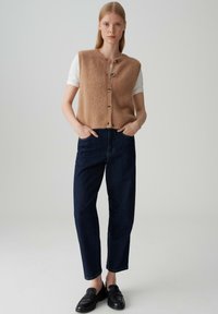 Gilet lavorato a maglia beige e morbido con scollatura rotonda e chiusura con bottoni, indossato sopra una camicia bianca. Abbinato a jeans a gamba dritta blu scuro e mocassini neri.