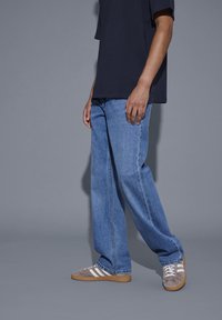 Person står iført løstsiddende blå jeans, grå sneakers med hvide striber og en mørk kortærmet skjorte mod en ensfarvet grå baggrund.