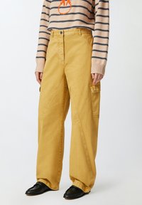 Žute široke hlače s prednjim džepom, s teksturiranim završetkom i dijagonalnim šavovima. Uparen s prugastim džemperom i crnim cipelama.