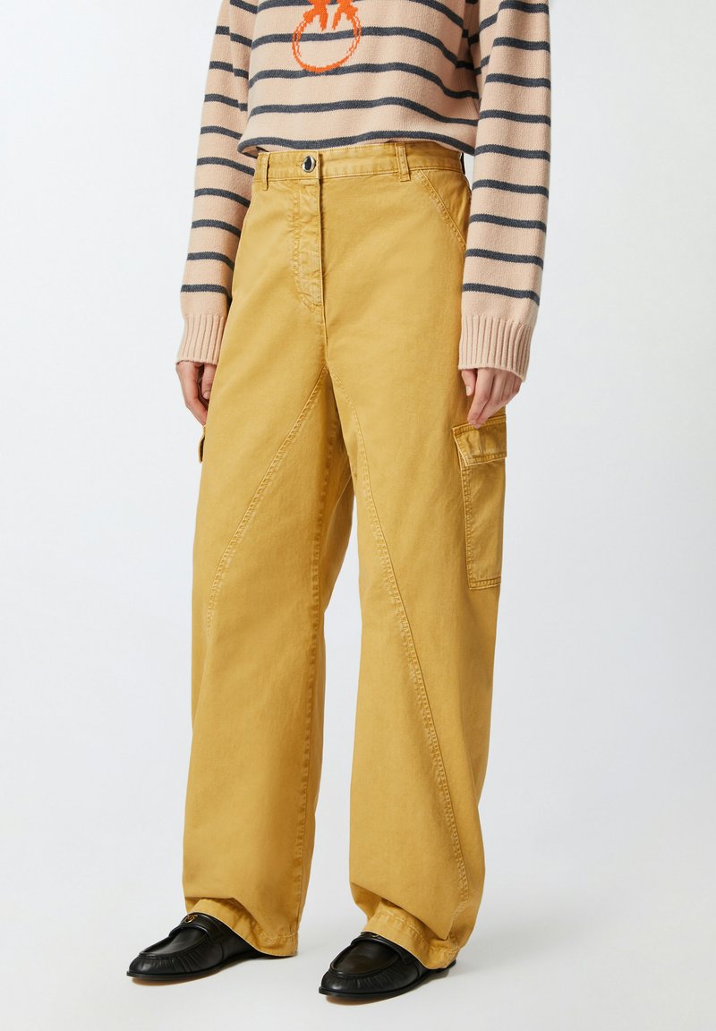 Žute široke hlače s prednjim džepom, s teksturiranim završetkom i dijagonalnim šavovima. Uparen s prugastim džemperom i crnim cipelama.