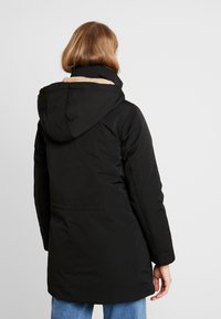 Parka noire matelassée avec capuche bordée de fourrure, dotée d'une fermeture éclair et de cordons ajustables à la taille. Manches longues avec poignets ajustés.