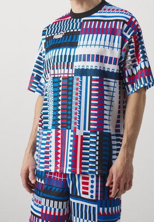 Mężczyzna ubrany w pasującą koszulę z krótkim rękawem i szorty z wyrazistymi geometrycznymi wzorami w czerwonym, niebieskim, czarnym i białym kolorze, z logo Puma.