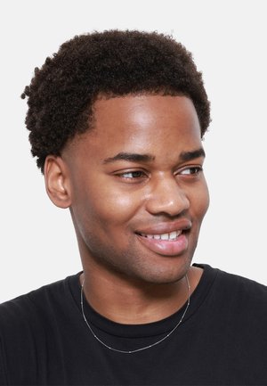 T-shirt noir à col rond, collier en chaîne argentée avec de petites accents ronds, cheveux courts et bouclés, souriant, fond neutre.