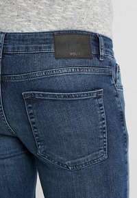 Jeans de mezclilla azul oscuro con un parche trasero de cuero negro con texto en relieve, dos bolsillos traseros y suaves detalles de deslavado.