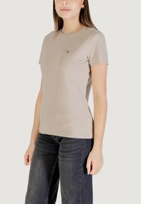 T-shirt beige a maniche corte realizzato in tessuto morbido con scollo a girocollo e un piccolo logo triangolare sul petto. Abbinato a jeans scuri.