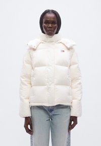 Κρεμ μπουφάν με υψηλό γιακά, μεγάλες ραμμένες ενότητες και λογότυπο στην αριστερή πλευρά του στήθους. Διαθέτει κουμπιά με πατητά κλιπ στον γιακά.