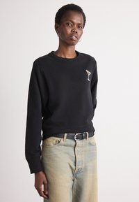 Sudadera negra con cuello redondo y mangas largas, que presenta un pequeño diseño de una copa de martini embellida en el pecho. Combinada con jeans de color azul claro.