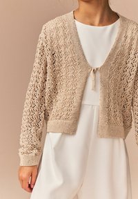 Beige strikket cardigan med åben front og indviklet blonde mønster. Har ribbede ærmekanter og bindebånd ved halsudskæringen.