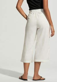 Fehér vászon culottok, illeszkedő, elasztikus derékkal, rövid, bő lábakkal és puha textúrával. Fekete szandálokkal párosítva.