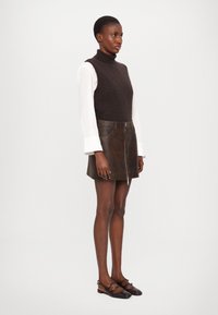 Maglione collo alto in lana marrone sopra una camicia a maniche lunghe bianca, abbinato a una mini gonna in pelle marrone con zip e scarpe basse scure.