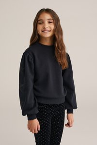 Tmavomodrá mikina s nafouklými rukávy, zdobená stříbrnými hvězdnými vzory na rukávech. Spárovaná s černými sametovými kalhotami s puntíky.