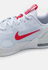 Chaussure de sport blanche avec un swoosh Nike rouge, lacets blancs, languette avec onglet rouge et empeigne en mesh texturé pour respirabilité et soutien.