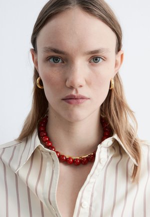 Mujer joven con cabello castaño claro que lleva pendientes de aro dorados, un collar de cuentas rojas y una camisa de botones a rayas crema sobre un fondo blanco.