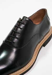 Chaussure habillée en cuir noir avec un bout arrondi, un design à lacets, une semelle intermédiaire contrastante couleur tan et une semelle extérieure en caoutchouc texturé.
