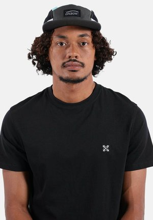 Homme aux cheveux bouclés portant une casquette Oxbow noire et un t-shirt noir avec un petit logo blanc sur la poitrine, faisant face à l'avant sur un fond blanc.