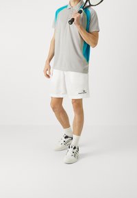 Šedé polo tričko s modrými a zelenými akcenty, spárované s bílými sportovními šortkami se logem. Model drží tenisovou raketu a má na sobě sportovní boty.