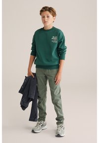 Zelena blagovna majica s tekstom na prsih, kombinirana s svetlozelenimi hlačami. Model drži temno modro jakno. Tenisice so sivo z črnimi detajli.