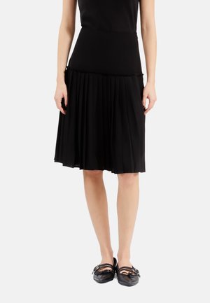 Woman wearing a black sleeveless top, knee-length pleated black skirt, and black flat shoes with straps, standing on a white background.