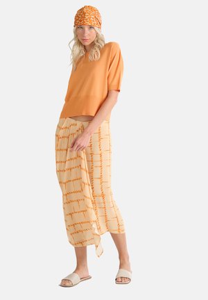 Haut tricoté orange à manches courtes, associé à une jupe jaune à motif avec des lignes oranges. Le mannequin porte des sandales à glisser beiges et un foulard orange.