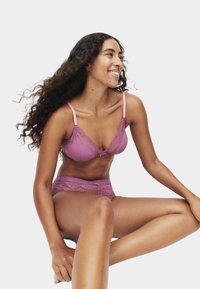 Kobieta z długimi kręconymi włosami siedzi i uśmiecha się, ubrana w dopasowany fioletowy koronkowy komplet bielizny na jednolitym białym tle.