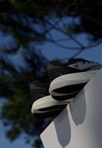 Svartvita Adidas-sneakers som vilar på en vit yta utomhus med suddig grön lövverk och blå himmel i bakgrunden.