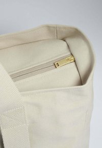 Interiér béžové plátěné tašky ukazuje kapsu na zip se zlatým táhlem, hladkou texturu a pevně šité švy.