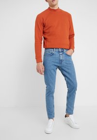 Pomarańczowa krótka bluza, niebieskie zwężane jeansy i białe trampki. Gładka tkanina z prążkowanym dekoltem i mankietami na rękawach.