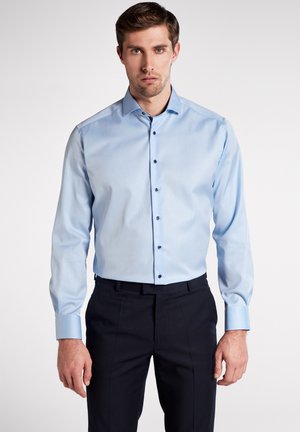 Hellblaues Langarmshirt mit klassischem Kragen, Knopfleiste und schwarzen Knöpfen, kombiniert mit dunkelgrauen Hosen. Glatte Stoffstruktur.