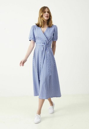 Kvinde iført hvide sneakers og en blå- og hvidternet omslagskjole med korte pufærmer, gående mod en ensartet lys baggrund.