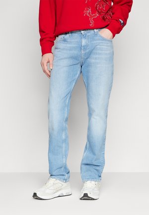 Mężczyzna ubrany w jasnoniebieskie dżinsy, białe trampki i czerwony sweatshirt z motywem smoka, stoi z jedną ręką w kieszeni.