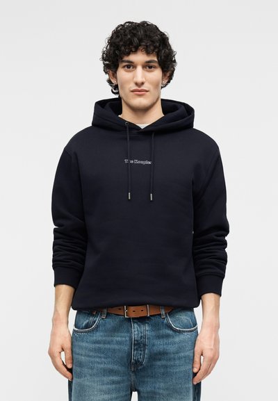 Jeune homme aux cheveux bouclés portant un sweat à capuche noir avec cordons de serrage et un jean bleu, sur un fond uni.