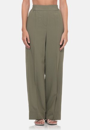 Donna con pantaloni a vita alta e gamba larga verde oliva e sandali bianchi aperti, in piedi davanti a uno sfondo chiaro e uniforme.