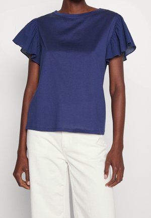 Ciemnoniebieska bluzka z krótkim rękawem i falbankami na rękawach, zestawiona z białymi, wysokim stanem spodniami na stojącej osobie.