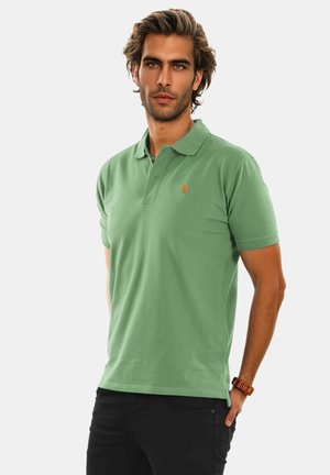 Mann mit hellbraunen Haaren, der ein grünes Poloshirt und schwarze Hosen trägt, steht mit einer Hand in der Hosentasche vor einem einfarbigen Hintergrund.