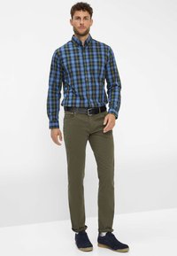 Chemise à carreaux bleue, verte et noire ; associée à un pantalon vert olive. Comprend une ceinture noire et des baskets navy.