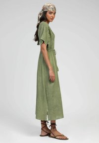 Robe verte, longueur cheville, avec des manches courtes à volants, taille cintrée et fentes sur les côtés. Présente une texture douce et une coupe décontractée. Portée avec des sandales.