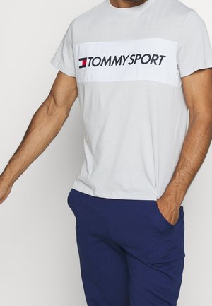 Mand iført en hvid Tommy Sport t-shirt og blå bukser med den ene hånd i lommen mod en ensfarvet baggrund.