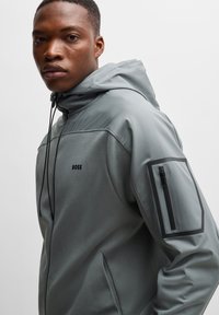 Jongeman in een grijze hooded jack met een ritszak op de arm en het "BOSS"-logo op de borst, recht vooruit kijkend tegen een effen achtergrond.
