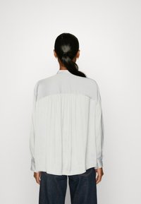 Camicia grigia chiaro con una texture liscia, dotata di uno schienale plissettato, maniche lunghe e collo a camicia, abbinata a jeans blu scuro.