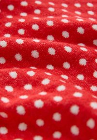 Punainen kangas valkoisilla pilkuilla, teksturoitu pinta, pehmeä materiaali, laskostettu näkyvin taittein ja kevyillä varjoilla, jotka korostavat kuviota.