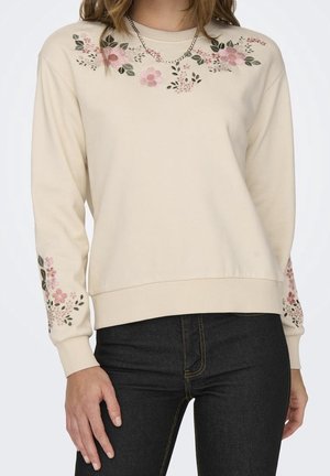 Cream sweater with pink floral embroidery on chest and sleeves, worn with dark jeans and a silver chain necklace.