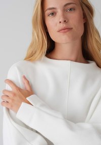 White, lightweight sweater with a wide neckline, long sleeves, and a textured finish. Features a subtle vertical seam at the center.