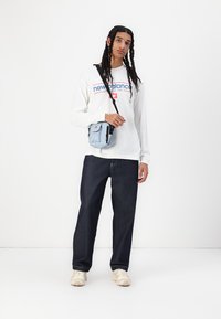 Hvit langermet skjorte med blå og rød "New Balance"-logo, mørkeblå jeans, beige sneakers og en liten lyseblå crossover-veske.