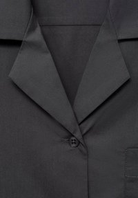 Camisa negra con cuello, que presenta una textura suave, cierre de botones y un bolsillo. Las solapas afiladas crean un diseño estructurado.