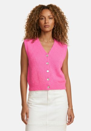 Femme aux cheveux bouclés portant un gilet sans manches rose vif à boutons sur le devant et une jupe blanche, debout devant un fond uni et clair.