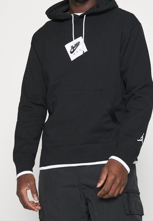 Hoodie noir avec poche kangourou, cordons blancs et un patch logo sur la poitrine. Poignets côtelés avec accents blancs.