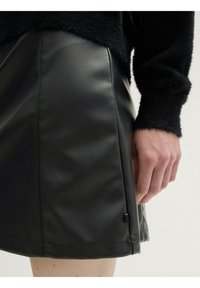 Mini falda de piel sintética negra con una textura suave, que presenta costuras verticales y una pequeña etiqueta de acento en el lado. Mano posicionada cerca del dobladillo.