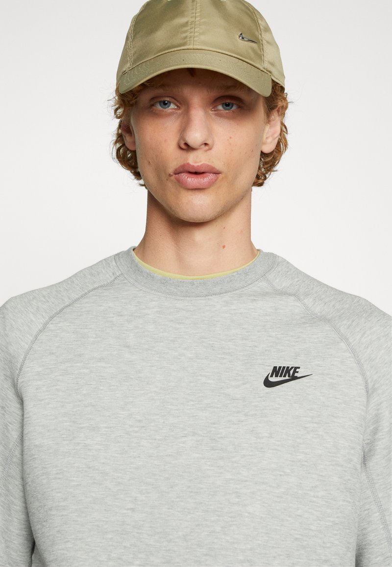 Sweatshirt de sport gris clair avec un col rond, arborant un logo Nike noir sur la poitrine. Porté avec une casquette kaki.
