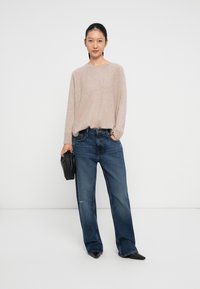 Light beige sweater with long sleeves, paired with relaxed-fit blue jeans. Model holds a black handbag, wearing pointed black shoes.