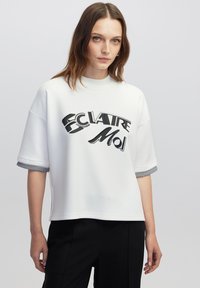 Žena se rovnými hnědými vlasy, oblečená v bílé košili s krátkým rukávem se textem "ÉCLAIRE Moi" a černými kalhotami, na jednobarevném pozadí.